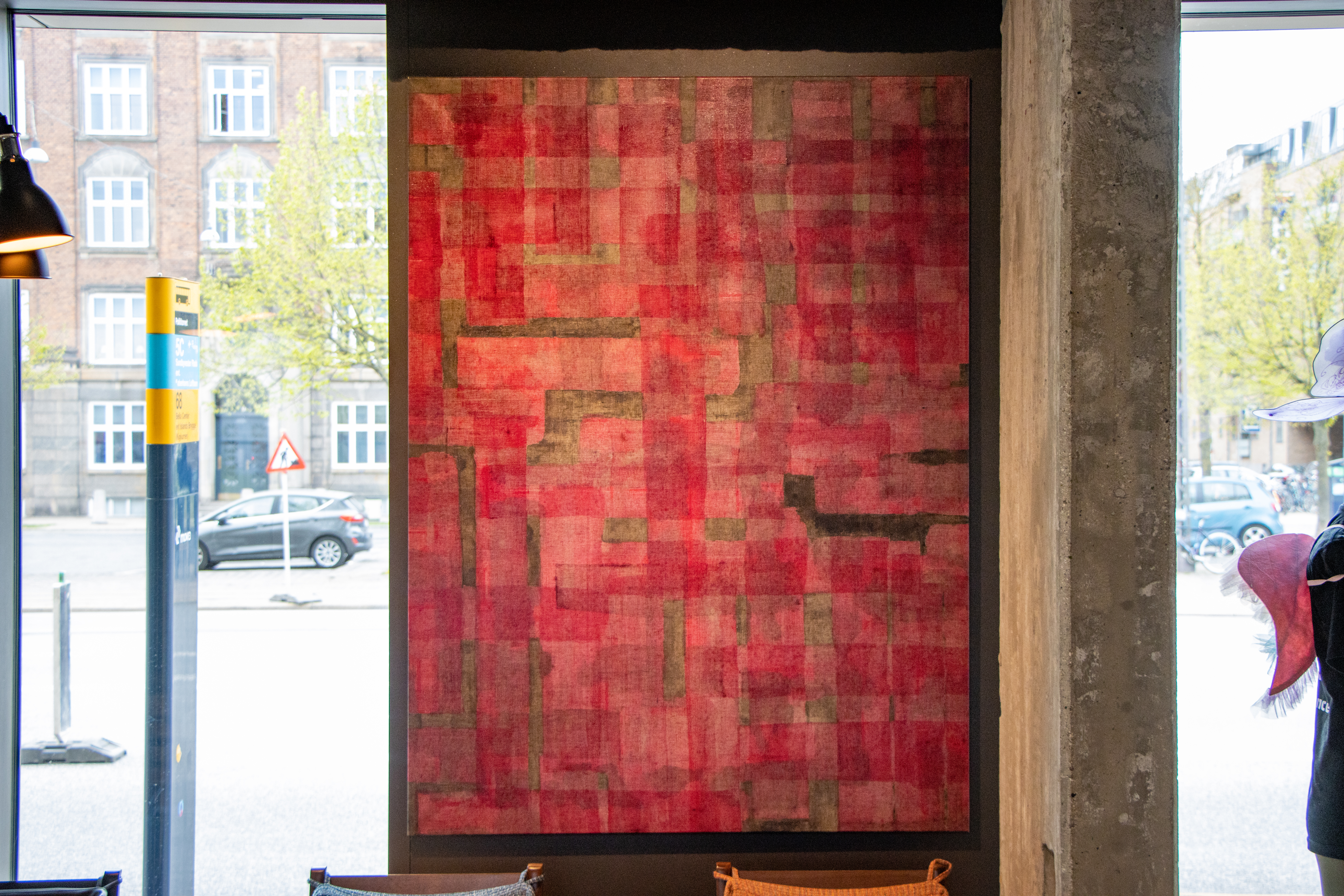 A large abstract painting with a red and pink geometric pattern, accented by occasional dark lines and rectangles. It hangs on a wall beside a large window overlooking a street with parked cars and brick buildings.